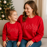 Woman and child wearing red 'Merry' sweatshirts sitting on a couch with a Christmas tree in the background.